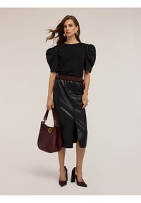 Nainen, jolla on päällään musta puffihihainen paita ja mustat nahkahameet, pitää burgundin väristä käsilaukkua, jalassaan burgundin väriset korot, seisoo yksinkertaista taustaa vasten.