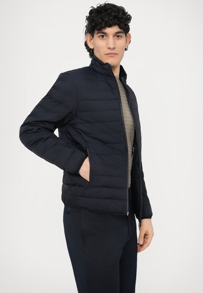 Temno modra podložena jakna z ovratnikom, žepi z zadrgo spredaj in teksturirano tkanino. Nošena čez svetlo rjav vzorčast zgornji del.