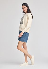 Felpa crema con dettagli del logo neri, abbinata a una gonna di jeans blu e sneakers avorio. Tessuto morbido, vestibilità casual e accenti semplici.