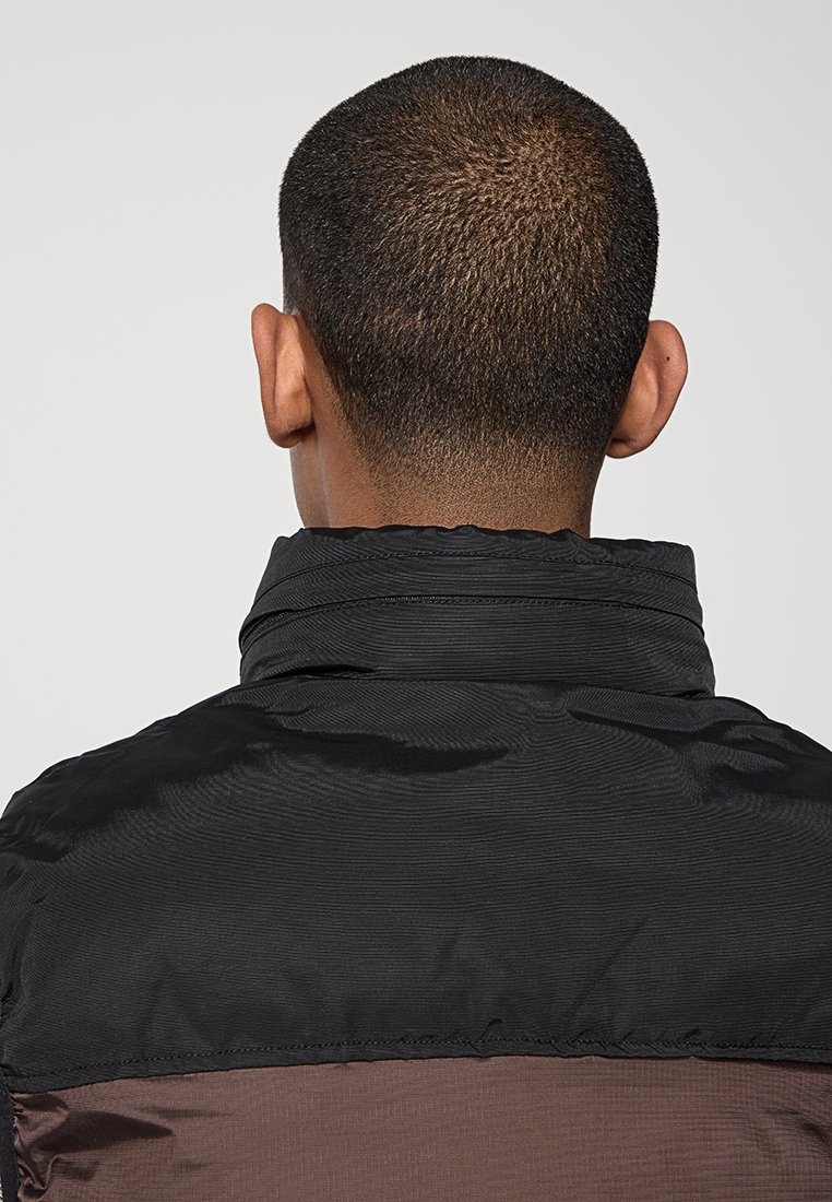 Personne avec des cheveux courts et foncés vue de derrière, portant une veste noire et marron avec un col relevé sur un fond uni.