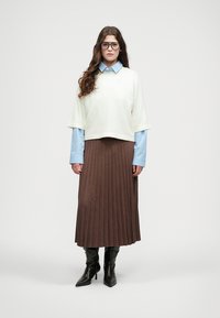 Light, cropped sweater in cream over a blue striped button-up shirt, paired with a long, pleated brown skirt and black knee-high boots.