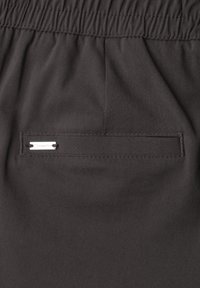 Black pants fabric with a horizontal back pocket featuring a small white rectangular label stitched on the pocket edge.