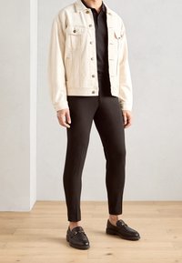 Světle béžová džínová bunda s předními kapsami na černém polo tričku, doplněná o černé slim-fit kalhoty a černé mokasíny se zlatými akcenty.