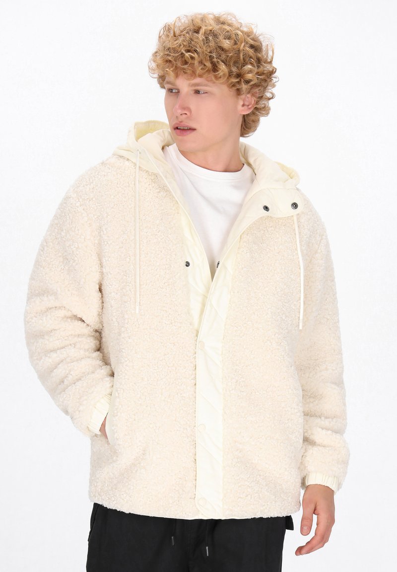 Sherpa-jakke i creme med foret hætte, quiltede detaljer, elastiske manchetter og trykknaplukninger. Blød tekstur med et hyggeligt design.