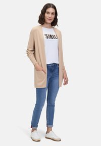Beige lange cardigan met open voorkant en lange mouwen, gecombineerd met een wit graphic t-shirt en blauwe skinny jeans. Schoeisel: witte sneakers.
