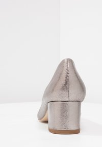 Chaussure à talon argenté métallisé avec un bout rond et un petit talon bloc. La surface présente une finition texturée et un détail de couture sans couture.