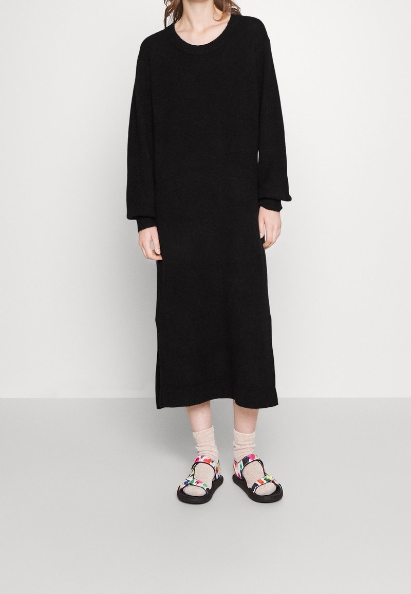 Personne portant une longue robe en maille noire avec des chaussettes beiges et des sandales noires à brides colorées, debout devant un fond uni clair.