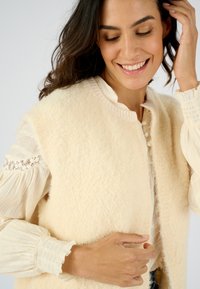 Chaleco de lana marfil con un acabado suave y texturizado, con cuello redondo y cierre de botones, usado sobre una blusa de crema con volantes y adornos de encaje.