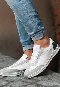 Weiße und graue Sneaker mit einem Obermaterial aus Leder und Wildleder, perforierten Details und einem strukturierten schwarzen Akzent an der Ferse. Die Sohle ist weiß.