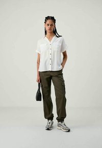 Camicia bianca con maniche corte e due tasche sul petto, abbinata a pantaloni cargo verde oliva. Gli accessori includono una piccola borsa nera.