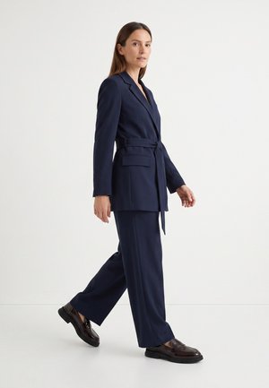 Frau läuft und trägt einen marineblauen Blazer mit Gürtel, passende weite Hosen und dunkelbraune Loafers vor weißem Hintergrund.