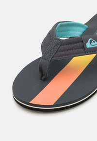 Dunkelgraue Flip-Flops mit strukturiertem Fußbett, ausgestattet mit einem türkisfarbenen Riemen und einem mehrfarbigen diagonalen Streifen in Orange und Gelb.