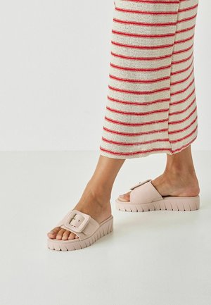 Pink platform slide sandals with a wide strap featuring a decorative buckle. Textured, ridged sole for grip. Worn with a striped knit skirt.