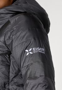 Melnā izolētā jaka ar teksturētu virsmu, ar kapuci un Eiger Extreme logotipu kreisajā piedurknē. Viegls un kompakts dizains.