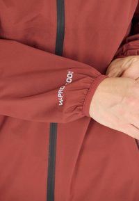 Röd vattentät jacka med hög krage, elastiska ärmslut, svart dragkedja och "W-PRO 5000"-logga på ärmen. Slät textur.