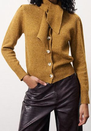 Frau trägt eine senfgelbe, geknöpfte Strickjacke mit einem gebundenen Halstuch und dunkelbraune, strukturierte Lederhose, Hand in der Tasche.