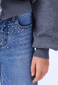 Pull en tricot gris avec des poignets côtelés, associé à un jean en denim bleu, présentant des embellissements brillants éparpillés sur l'avant et la zone des poches.
