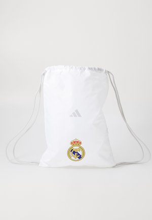 Mochila blanca con cordón presenta el logo de Adidas y el emblema del Real Madrid en la parte frontal, contra un fondo minimalista.