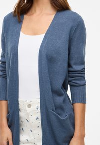 Μπλε πλεκτό cardigan με σχέδιο V-neck, ριμπ μανσέτες και δύο μπροστινές τσέπες, σε συνδυασμό με ένα λευκό τοπ και patterned παντελόνι.