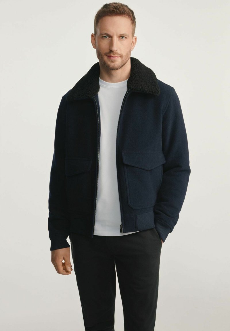 Mężczyzna ubrany w granatową kurtkę z czarnym futrzanym kołnierzem, białą koszulkę i czarne spodnie, stojący na tle jasnego, jednolitego tła.