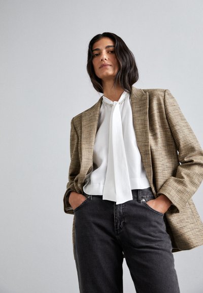 Brunternet blazer over en hvid bluse med en slipsdetalje, parret med mørkegrå jeans. Glatte teksturer og en neutral farvepalette.