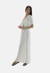 Hvid maxi-kjole med pufærmer og smocked bodice, der har en fronttie-detalje. Modellen bærer sorte sandaler med geometriske stropper.