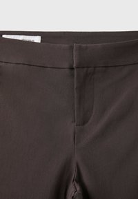 Gros plan de la ceinture et de la couture avant d'un pantalon marron foncé avec une étiquette visible à l'intérieur sur fond blanc.