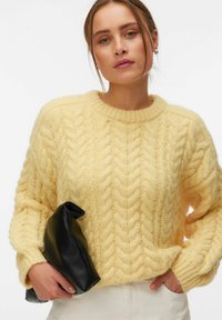 Femme portant un pull en maille torsadée jaune tenant une pochette en cuir noire, avec un pantalon blanc et un maquillage neutre.