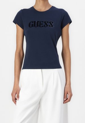 Granatowy top z krótkim rękawem z czarnym, ćwiekowanym logo "GUESS" zestawiony z białymi, szerokimi spodniami z wysokim stanem na stojącej osobie.