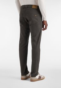 Moški, ki nosi temno karirane hlače in bele športne copate z rjavimi poudarki, stoji ob brezhibni beli podlagi, prikazan od zadaj.