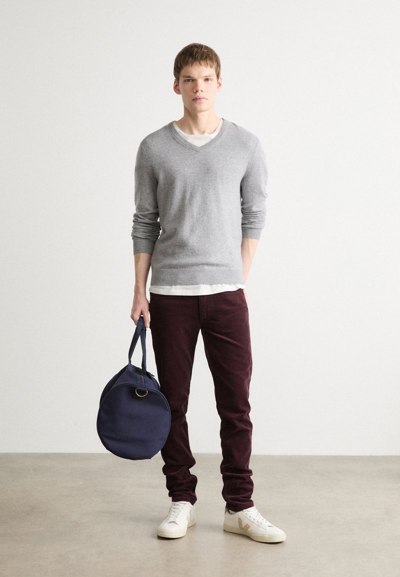 Grauer Woll-V-Ausschnitt-Pullover über einem weißen T-Shirt, bordeauxfarbene Cordhose, cremefarbene Sneakers und eine runde marineblaue Tasche mit Griff.