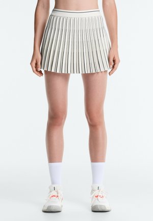 Kvinde iført hvid plisseret nederdel med tynde sorte striber, hvide sokker og hvide sneakers, stående mod en ensfarvet lys baggrund.