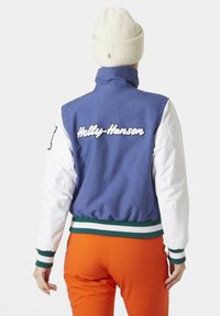 Blauwe en witte bomberjack met ribgebreide boorden en zoom, met "Helly Hansen" geborduurd op de achterkant in het wit. Gecombineerd met oranje broek.