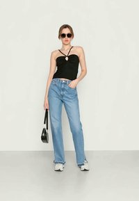 Top preto sem mangas com um detalhe de buraco na parte da frente, combinado com jeans de cintura alta azul claro e ténis brancos. Acessorizado com uma pequena mala preta.