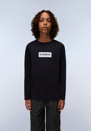 Zwarte lange mouwen T-shirt van katoen, met een witte rechthoekige logo in het midden. G gedragen met donkere cargobroek, staande tegen een lichte achtergrond.