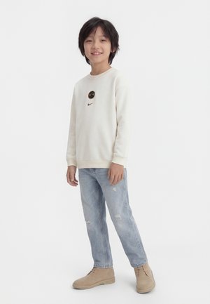 Garçon souriant debout, portant un sweat-shirt blanc avec un logo, un jean bleu clair déchiré et des bottines beige sur fond blanc.