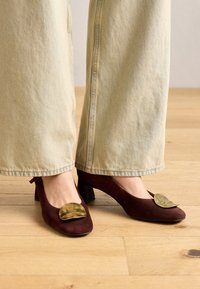 Scarpe con tacco in suede bordeaux con punta arrotondata e un accento metallico dorato sulla parte superiore, abbinate a jeans a gamba larga blu chiaro.