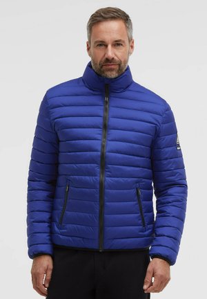 Man with short hair and beard wearing a blue quilted jacket with zipped pockets and a stand-up collar against a plain background.