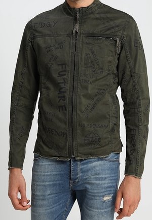 Mann trägt eine dunkelgrüne Jacke mit Reißverschluss, bedruckt mit schwarzen, handgeschriebenen Worten wie "ZUKUNFT", "FREIHEIT" und "FRIEDEN", kombiniert mit blauen, zerrissenen Jeans.