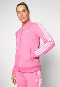 Rozā adidas jaka ar rāvējslēdzēju, kurai ir baltas trīs svītras uz piedurkniem, ar stāvošu apkakli un sānu rāvējslēdzēju kabatām. Mīksta, gluda auduma.