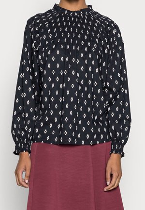 Blouse noire manches longues avec motif ovale blanc, col montant froncé et poignets, portée avec une jupe bordeaux.