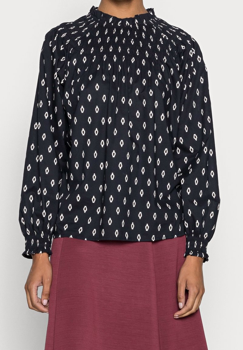 Blouse noire manches longues avec motif ovale blanc, col montant froncé et poignets, portée avec une jupe bordeaux.
