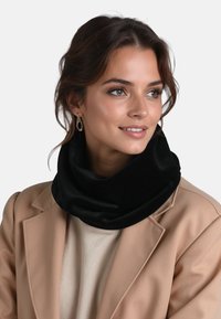 Mujer sonriente, con abrigo beige, suéter crema, cuello de terciopelo negro y pendientes dorados colgantes, mirando ligeramente hacia la derecha sobre un fondo liso.