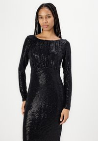 Robe longue à manches longues en sequins noirs avec un décolleté bateau, une silhouette cintrée et un détail de corsage froncé. La surface présente un motif texturé et brillant.