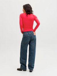 Kvinne med langt mørkt hår iført en rød langermet skjorte, vide blå jeans og svarte sko, stående med hendene på hoftene og vendt bort.