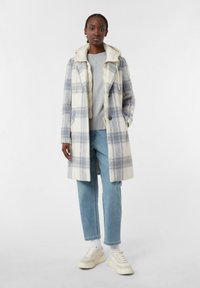 Cappotto a quadri in morbido tessuto con cappuccio, caratterizzato da tonalità di blu e bianco, due tasche anteriori e bottoni. Abbinato a jeans azzurri e sneakers.