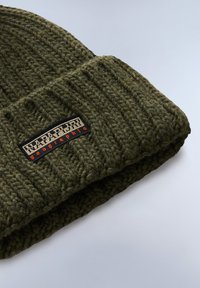 Gorro de punto color verde oliva con textura acanalada y ala plegable. Presenta un parche rectangular con "NAPAPIJRI" y "geographic" en colores contrastantes.