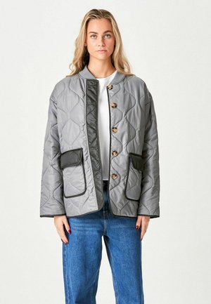 Jonge vrouw met lang blond haar draagt een grijze doorgestikte jas met grote knopen, zakken met zwarte rand, wit shirt en blauwe jeans.