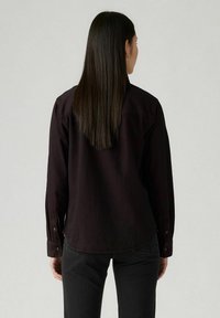 Chemise noire à manches longues en tissu lisse, avec un col et un ourlet arrondi. La vue de dos met en avant une coupe décontractée.