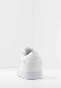 Vit sneaker med en slät syntetisk ovandel, vadderad krage, präglad logotyp på hälen och en gummisula med ett texturerat mönster.